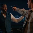 Ein Mann mit erhobenen Händen, bedroht von einem anderen Mann mit einer Pistole, in einer nächtlichen Stadtkulisse.