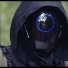Eine vermummte Figur mit dunklem Gewand und einem futuristischen Helm. Reflektierender Kreis mit blauen Lichtern sichtbar.