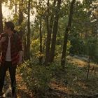 Eine Person mit rotem Hemd und Rucksack steht im sonnenbeschienenen Wald, umgeben von Bäumen und grünem Laub.