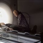 Eine Frau liegt in einem MRT-Scanner, während ein älterer Mann besorgt auf sie herabblickt.