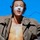 Ein Mann mit offenen Armen im Freien, trägt Mantel, nackter Oberkörper, weiße Gesichtsfarbe, blauer Himmel.