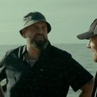 Zwei Männer unterhalten sich am Meer. Einer trägt einen Anglerhut, der andere eine Basecap. Der Hintergrund zeigt Wasser.