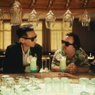 Zwei Männer in Sonnenbrillen sitzen an einer Bar mit exotischen Cocktails. Über ihnen hängen Gläser kopfüber.