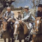Mehrere Männer in Cowboy-Kleidung reiten auf Pferden in einer trockenen, felsigen Landschaft.