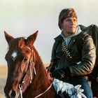 Ein Mann mit einer Lederjacke auf einem Pferd in einer offenen Landschaft. Er trägt einen großen Rucksack.