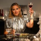 Eine Person in silbernem Kleid hebt feierlich ein Weinglas bei einem elegant gedeckten Tisch.