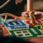 Ein kleiner, stehender Hamster auf einem Mensch-ärgere-dich-nicht-Spielbrett mit bunten Spielfiguren und einem Würfel.