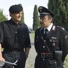 Ein Mann in Schwarz neben einem Polizisten in Uniform auf einem Weg, umgeben von Bäumen.