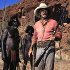 Es gibt keinen Text zu beschreiben. Ein Mann mit Cowboyhut hält ein Gewehr, umgeben von Männern mit Speeren in einer Wüstenlandschaft.