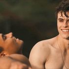 Zwei junge Männer ohne Hemd lächeln. Der eine liegt entspannt, der andere sitzt und schaut ihn freundlich an.