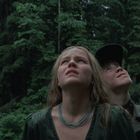 Zwei Menschen schauen mit besorgtem Gesichtsausdruck nach oben. Im Hintergrund ist ein dichter Wald zu sehen.