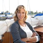 Eine Frau mit lockigem Haar steht lächelnd auf einem Segelboot, Boote und Wasser im Hintergrund.