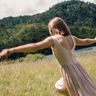 Eine Frau in einem hellen Kleid läuft über eine Wiese, Arme ausgestreckt, vor einem bewaldeten Hügel.