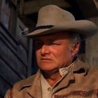 Ein Mann mit Cowboyhut steht vor einem Holzgebäude und schaut ernst.