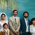 Eine Gruppe von sechs Erwachsenen und einem Kind posiert vor einem Hintergrund mit Palmen. Eine Frau trägt ein Hochzeitskleid.