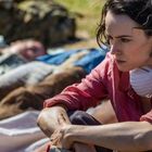 Eine Person mit ernstem Blick trägt eine Atemschutzmaske um den Hals und sitzt im Freien neben schlafenden Menschen.