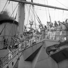 Viele Männer in Uniformen stehen auf einem Schiffdeck, jubeln und winken. Ein Schornstein im Hintergrund raucht.