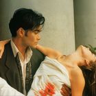 Ein Mann in einem Anzug hält eine Frau mit einem blutbefleckten weißen Kleid vor einem Pfeiler und Pflanzen.