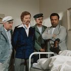 Vier Männer in verschiedenen Anzügen betrachten ein Krankenhausbett, auf dem jemand liegt.