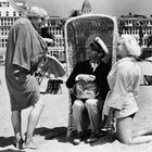 Person am Strand in hellem Mantel kniet vor einem Mann im Seemannsoutfit, der in einem Korbstuhl sitzt.