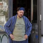 Ein Mann mit blauem Stirnband trägt ein T-Shirt mit dem Text: "Was wird Yvonne dazu sagen?" und einen gestreiften Bademantel.