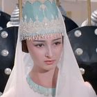 Keine sichtbaren Texte. Eine Frau in traditioneller Kleidung mit einer verzierten Haube vor schwarz-silbernen Schilden.