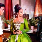 Drei Personen in historischen Kostümen stehen in einem Raum. Eine Frau in grünem Kleid mit aufwendiger Frisur im Zentrum.