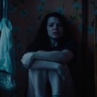 Eine Person sitzt ängstlich auf dem Boden eines blauen Zimmers, umgeben von einem Bett, Spiegel und Kommode.