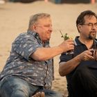 Zwei Männer sitzen barfuß am Strand, entspannen und trinken Cocktails aus Gläsern, einer aus Kupfer.