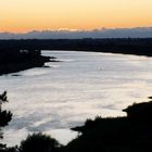 Keine Texte sichtbar. Ein Fluss fließt durch eine Landschaft bei Sonnenuntergang, umgeben von Bäumen und Wiesen.