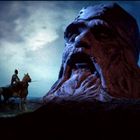 Ein Ritter auf einem Pferd schaut zu einem riesigen in Stein gemeißelten Kopf mit offenem Mund, vor einem düsteren, wolkigen Himmel.