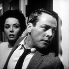 Es gibt keinen Text. Ein Mann in Anzug und Krawatte schaut angespannt durch eine Tür, eine Frau steht hinter ihm.