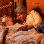 Eine Frau liegt neben einem schlafenden Kind in einem Holzbett. Sie trägt ein Kopftuch, das Kind hat blonde Zöpfe.