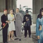 Fünf Personen stehen in einem Säulengang. Eine Frau im Vordergrund trägt einen glitzernden, blauen Anzug.