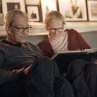 Zwei ältere Personen sitzen auf einem Sofa und betrachten gemeinsam ein großes Fotoalbum. Im Hintergrund sind gerahmte Fotos.