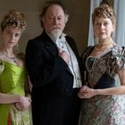 Drei Personen in historischer Kleidung, Mann in schwarzem Anzug, Frauen in grünen und gemusterten Kleidern mit Perlenschmuck.