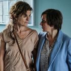 Zwei Personen stehen eng zusammen und blicken sich intensiv an. Eine trägt ein beiges Shirt, die andere einen blauen Blazer.