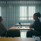 Zwei Personen sitzen sich in einem Raum mit Gitterstäben gegenüber. Ein Mikrofon steht zwischen ihnen auf dem Tisch.