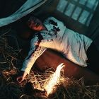 Ein Mann in blutbespritztem Hemd liegt in einem dunklen Raum auf Stroh, mit einer kleinen Flamme nahe seiner Hand.