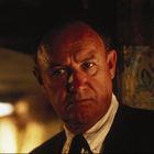 Ein Mann mit ernstem Gesichtsausdruck in einem schwach beleuchteten Raum, im Hintergrund leichte Unschärfen.