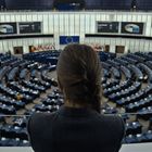 Zwei Monitore zeigen das Logo der EU. Eine Frau im Vordergrund blickt auf einen großen Konferenzsaal mit vielen Sitzreihen.