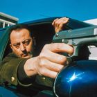 Ein Mann mit ernstem Blick zielt mit einer Pistole durch ein Autofenster. Im Hintergrund ein modernes Gebäude.