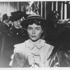 Eine Frau mit einem Strohhut und ernstem Gesichtsausdruck sitzt, umgeben von anderen Menschen im Hintergrund.