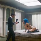 Ein junger Mann fotografiert eine Person mit blauen Haaren auf einem Bett in einem Raum mit großen Fenstern und Vorhängen.