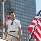 Ein Mann in weißem Polohemd hält ein Weinglas, steht auf einer Yacht mit US-Flagge, Hochhäuser im Hintergrund.