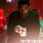 Ein Mann hält ein Glas in einer Bar mit rotem und grünem Licht, umgeben von anderen Personen im Hintergrund.