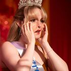 Eine junge Frau mit Krone hält erschrocken ihr Gesicht, trägt ein weiß-lila Band mit einer unleserlichen blauen Aufschrift.