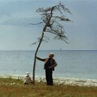 Ein älterer Mann steht neben einem kahlen Baum am Seeufer, ein Kind sitzt daneben im Gras.