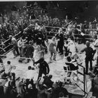 Im Mittelpunkt steht ein belebter Boxring mit vielen Menschen, einige Fotografen und ein Schiedsrichter.