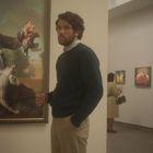 Ein Mann steht vor einem Gemälde eines Rokoko-Jägers mit Hund und Vogel. Hinter ihm betrachten zwei Personen andere Kunstwerke.